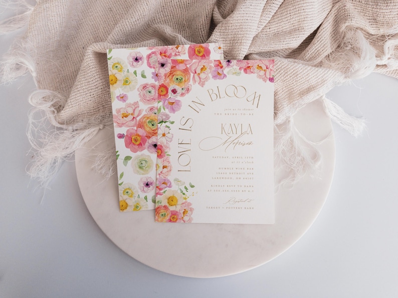 May include: Two wedding invitations with floral designs. The invitations feature a watercolour floral border in shades of pink, yellow, and white. The text on the invitation reads "Love is in Bloom" and includes the names "Kayla Melissa".