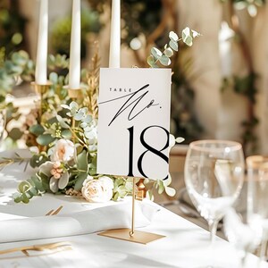 May include: White table number card with black calligraphy that says "Table No. 18" on a gold metal stand. The card is sitting on a white tablecloth with flowers and candles in the background.