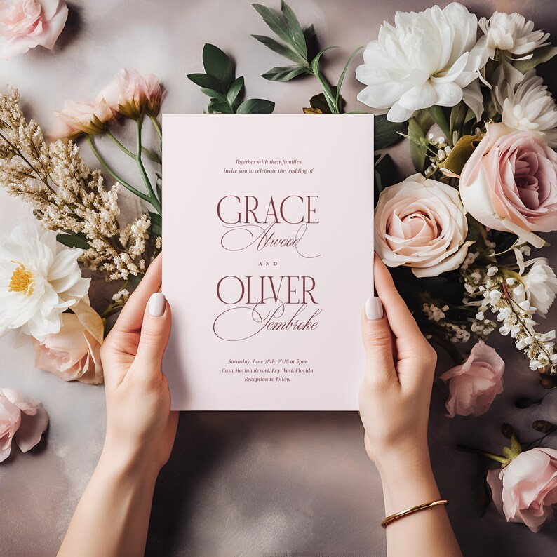 Puede incluir: Una invitaci&oacute;n de boda rosa claro con escritura elegante, sostenida por manos con una pulsera dorada. La invitaci&oacute;n presenta los nombres "Grace Atwood" y "Oliver Sembriche" con detalles florales, rodeada de flores rosas y blancas.