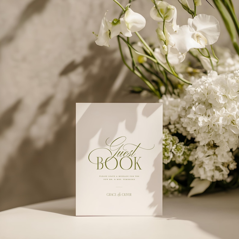 May include: A white guest book with the words "Guest Book" in elegant green script. The book also includes the text "Please leave a message for the new Mr. & Mrs. [surname]" and the name "Grace & Oliver". White flowers are in the background.