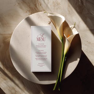 Puede incluir: Una tarjeta de men&uacute; de boda con el texto "The Menu" en escritura burdeos, colocada sobre un plato redondo y blanquecino. Dos calas blancas con tallos verdes descansan junto al men&uacute;. La escena est&aacute; iluminada con luz natural.