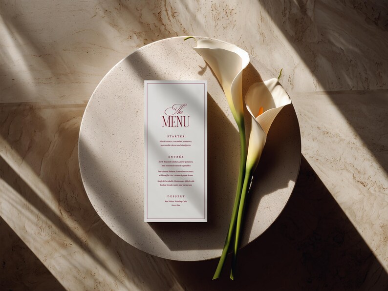 Puede incluir: Una tarjeta de men&uacute; de boda con el texto "The Menu" en escritura burdeos, colocada sobre un plato redondo y blanquecino. Dos calas blancas con tallos verdes descansan junto al men&uacute;. La escena est&aacute; iluminada con luz natural.