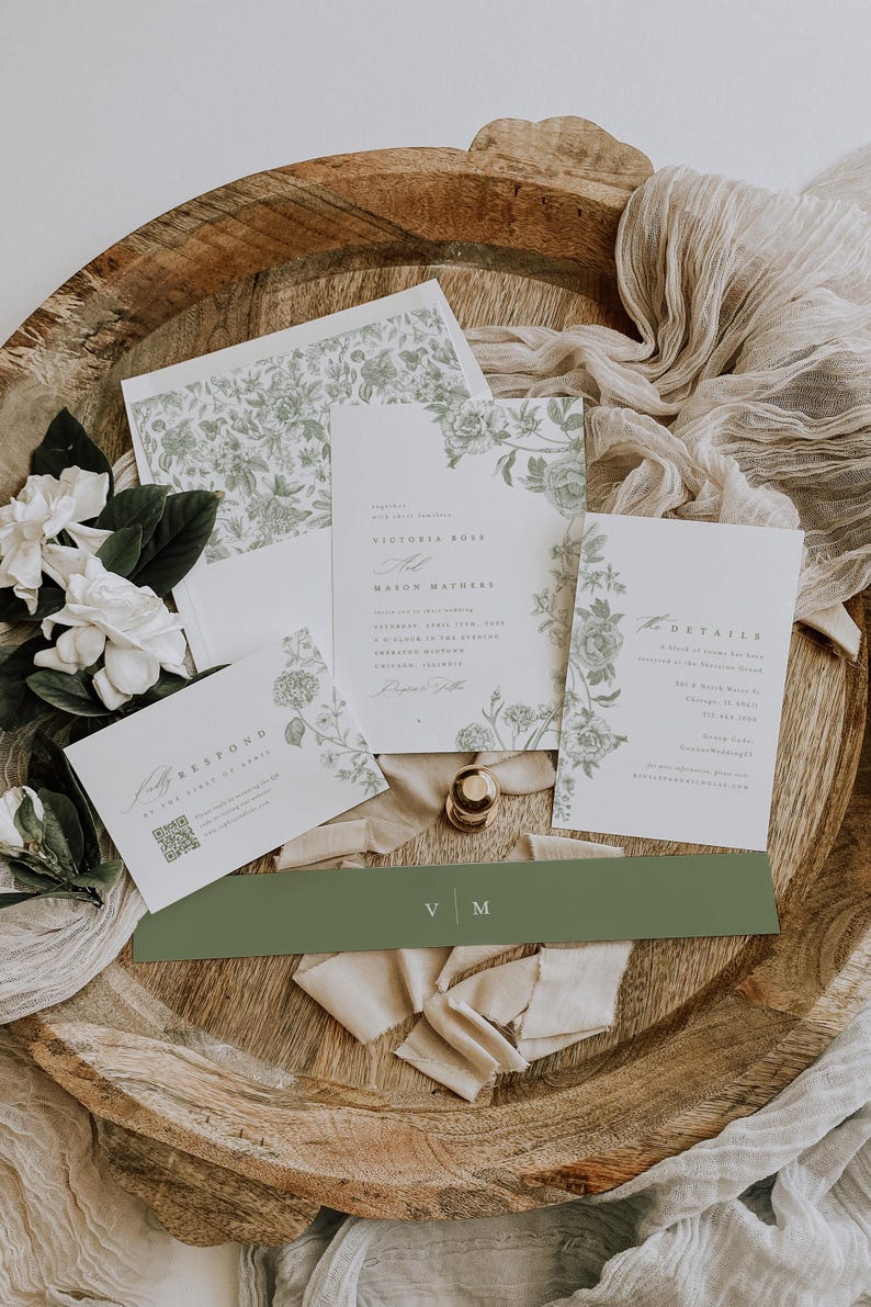May include: A wedding invitation suite featuring white paper cards with sage green floral illustrations. The set includes a main invitation, a details card, a response card with a QR code, and a solid sage green belly band with the initials V M.