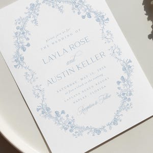May include: A wedding invitation with a blue floral border. The text reads "Please join us for the wedding of Layla Rose and Austin Keller, Saturday, May 15, 2026 at five o'clock in the evening, Laplaya Beach Resort, Naples, Florida. Reception to follow."