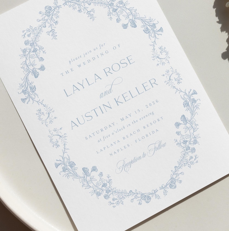 May include: A wedding invitation with a blue floral border. The text reads "Please join us for the wedding of Layla Rose and Austin Keller, Saturday, May 15, 2026 at five o'clock in the evening, Laplaya Beach Resort, Naples, Florida. Reception to follow."