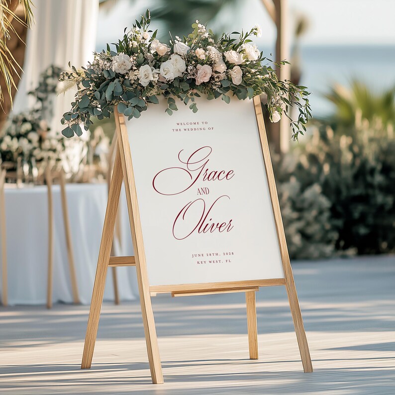 Puede incluir: Un caballete de madera con las palabras "Grace and Oliver" en escritura burdeos, adornado con un arreglo floral de rosas blancas y rosas y vegetaci&oacute;n. El letrero da la bienvenida a los invitados a una boda.