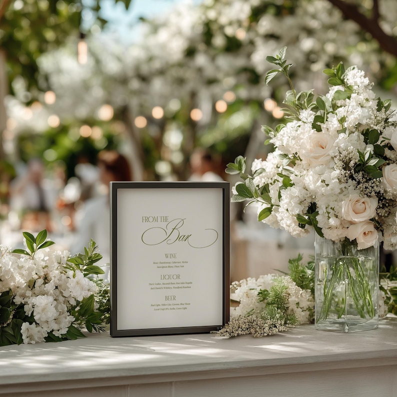 May include: A bar sign with the words "From the Bar" in elegant script, listing wine, liquor, and beer options. The sign is in a black frame, surrounded by white flowers and greenery, with a blurred background.