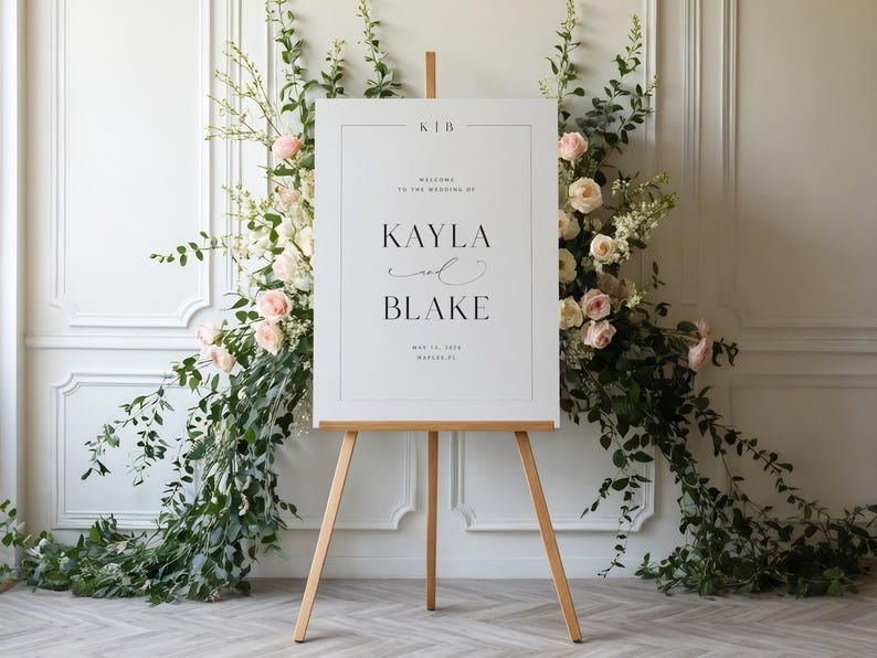 May include: A wedding welcome sign on a wooden easel, with the names Kayla and Blake in black script. The sign is surrounded by floral arrangements with pink and white roses and greenery.