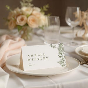 May include: A folded cream-coloured cardstock place card featuring the printed text AMELIA WESTLEY and table one, decorated with a green botanical floral illustration on the right side, positioned on a white ceramic dinner plate.