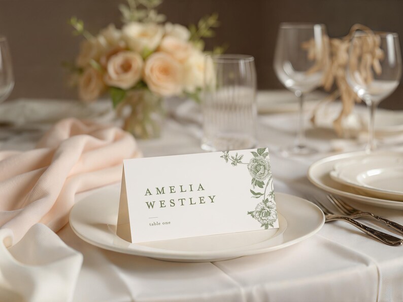 May include: A folded cream-coloured cardstock place card featuring the printed text AMELIA WESTLEY and table one, decorated with a green botanical floral illustration on the right side, positioned on a white ceramic dinner plate.