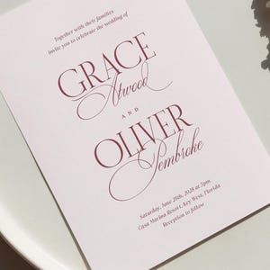 Puede incluir: Una invitaci&oacute;n de boda rosa claro con texto burdeos. La invitaci&oacute;n presenta los nombres "GRACE Atwood" y "OLIVER Yembreche". El texto tambi&eacute;n incluye la fecha, hora y lugar de la boda: Casa Marina Resort, Key West, Florida.