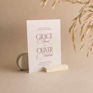 Puede incluir: Una tarjeta de invitaci&oacute;n de boda con los nombres "Grace Atwood" y "Oliver Jembicke" en escritura burdeos. La tarjeta se muestra sobre un bloque de madera y una peque&ntilde;a piedra redonda. Espigas de trigo secas en el fondo.