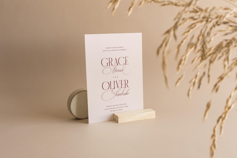 Puede incluir: Una tarjeta de invitaci&oacute;n de boda con los nombres "Grace Atwood" y "Oliver Jembicke" en escritura burdeos. La tarjeta se muestra sobre un bloque de madera y una peque&ntilde;a piedra redonda. Espigas de trigo secas en el fondo.