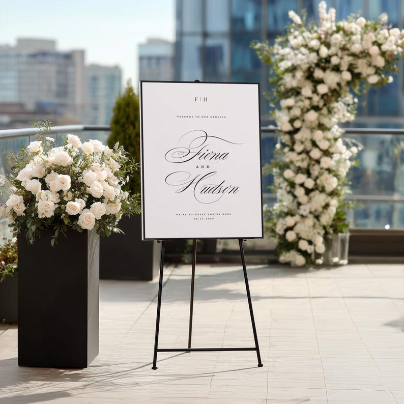 May include: A white rectangular wedding welcome sign with black text on a black metal tripod easel. The sign features the initials F|H, the names Fiona and Hudson in elegant script, and the date 09.17.2025. The sign is displayed outdoors.