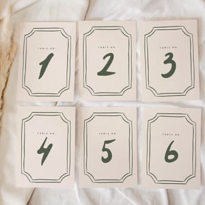 May include: Set of six table number cards. Each card features a cream background with a dark green border and number. The numbers 1 through 6 are displayed in a handwritten style. The words "TABLE NO." are printed at the top of each card.