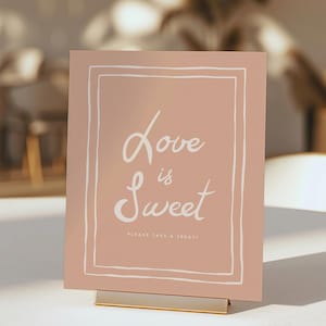 Könnte beinhalten: Ein rosa Schild mit weißem Rand und dem Text "Love is Sweet" in weißer Kursivschrift. Das Schild sagt auch "Please take a treat!" in kleinerer weißer Schrift.