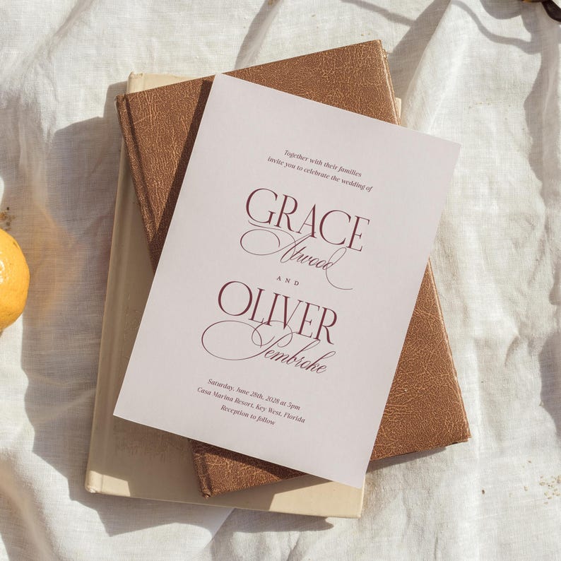 Puede incluir: Una invitaci&oacute;n de boda con los nombres "Grace Atwood" y "Oliver Sembiche" en escritura burdeos. La invitaci&oacute;n est&aacute; sobre un papel rosa claro, encima de dos libros marrones. El texto incluye la fecha y el lugar de la boda.