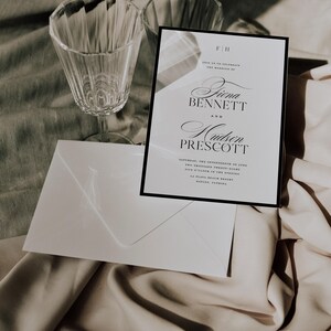May include: A wedding invitation with a black border, displaying the names 'Fiina Bennett' and 'Hudson Prescott' in script. Includes a white envelope and two crystal wine glasses. The invitation details the wedding date and location.
