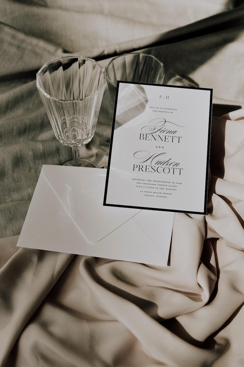 May include: A wedding invitation with a black border, displaying the names 'Fiina Bennett' and 'Hudson Prescott' in script. Includes a white envelope and two crystal wine glasses. The invitation details the wedding date and location.