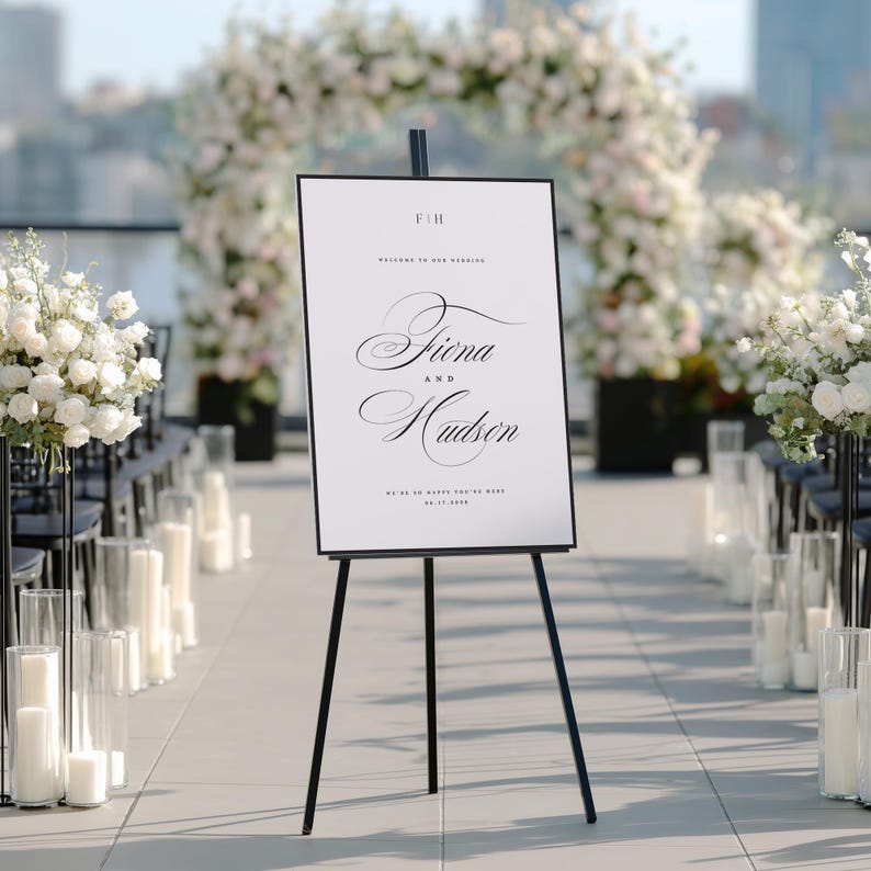 May include: A white rectangular wedding welcome sign with a thin black frame on a black tripod easel. The sign features the text F&H, Welcome to our wedding, Fiona and Hudson, We're so happy you're here, and the date 05.17.2025 in black font.