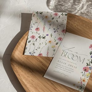 May include: Two floral-themed cards on a wooden tray. One card features a repeating pattern of pink, purple, and yellow flowers on a white background. The other card has the text "Love in Bloom" and event details, with floral accents.