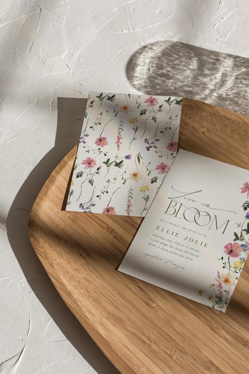May include: Two floral-themed cards on a wooden tray. One card features a repeating pattern of pink, purple, and yellow flowers on a white background. The other card has the text "Love in Bloom" and event details, with floral accents.