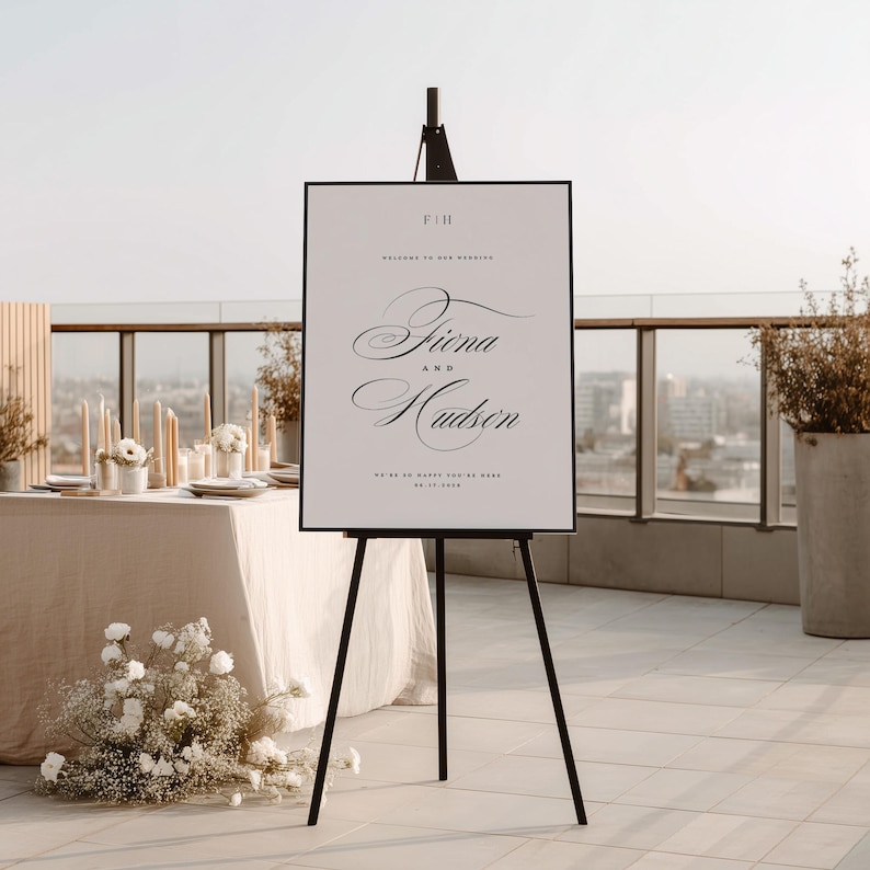 May include: A vertical rectangular wedding welcome sign on a black tripod easel. The white sign features black text reading F&H, Welcome to our wedding, Fiona and Hudson, and the date 10.17.2026 in a mix of serif and elegant script fonts.