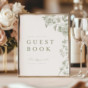 May include: A rectangular sign in a gold frame featuring the text PLEASE SIGN OUR GUEST BOOK in green, with the script text We're so happy you're here! and the names VICTORIA + MASON, decorated with green botanical floral illustrations on the right.
