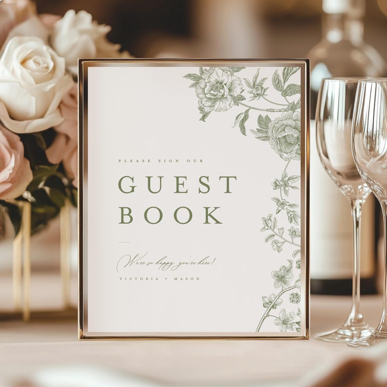 May include: A rectangular sign in a gold frame featuring the text PLEASE SIGN OUR GUEST BOOK in green, with the script text We're so happy you're here! and the names VICTORIA + MASON, decorated with green botanical floral illustrations on the right.