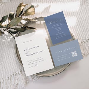 Op de afbeelding: Een huwelijksuitnodigingsset met een witte achtergrond en blauwe accenten. De uitnodigingskaart bevat de namen "Kinsley Moore" en "Nicholas Connor" in een eenvoudig, elegant lettertype. De RSVP-kaart bevat een QR-code waarmee gasten online kunnen reageren.