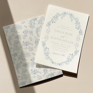 May include: A wedding invitation with a floral wreath design in blue on a white background. The invitation reads "Please join us for the wedding of Layla Rose and Austin Keller Saturday, May 13, 2023 at five o'clock in the evening Empire Place Brewery Naples, Florida Reception to follow"
