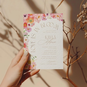 May include: A wedding invitation with the text "Love is in Bloom" and the names "Kayla & Anderson". The invitation features a watercolour floral design in pink, yellow, and purple hues. The invitation is held by a hand.