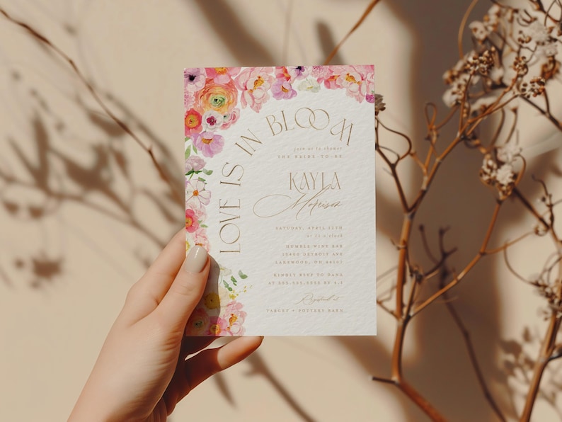 May include: A wedding invitation with the text "Love is in Bloom" and the names "Kayla & Anderson". The invitation features a watercolour floral design in pink, yellow, and purple hues. The invitation is held by a hand.