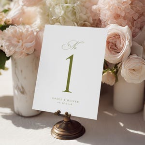 Pode incluir: Um cart&atilde;o branco com o n&uacute;mero da mesa "1" em verde oliva, exibido em um suporte de bronze. O cart&atilde;o tamb&eacute;m inclui os nomes "Grace & Oliver" e a data "06.28.2028". O cart&atilde;o est&aacute; situado em um fundo de flores rosa e brancas.
