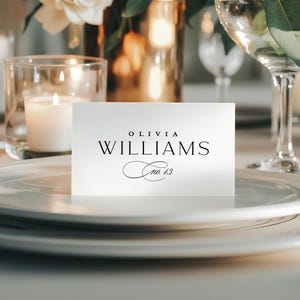Puede incluir: Una tarjeta de sitio blanca con el nombre "OLIVIA WILLIAMS" en escritura negra, con "no. 13" debajo. La tarjeta está sobre un plato blanco, con velas y cristalería en el fondo, creando una elegante mesa.