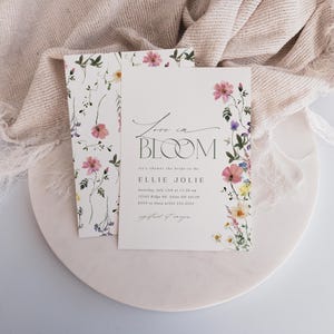 May include: A floral invitation suite featuring the words "Love in Bloom" in elegant script. The invitation is adorned with watercolor flowers in pink, yellow, and purple. A matching card with floral accents is also present, all on a white background.