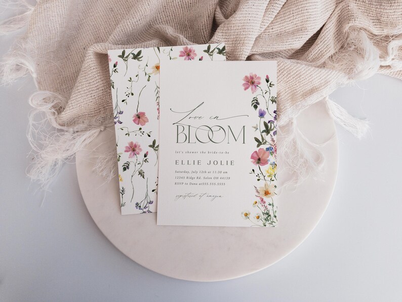 May include: A floral invitation suite featuring the words "Love in Bloom" in elegant script. The invitation is adorned with watercolor flowers in pink, yellow, and purple. A matching card with floral accents is also present, all on a white background.