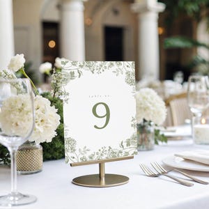May include: A table setting with a floral table number. The table number is white with a green floral border and the number 9. The table is set with white linens, glassware, and silverware. White flowers and candles are also present.
