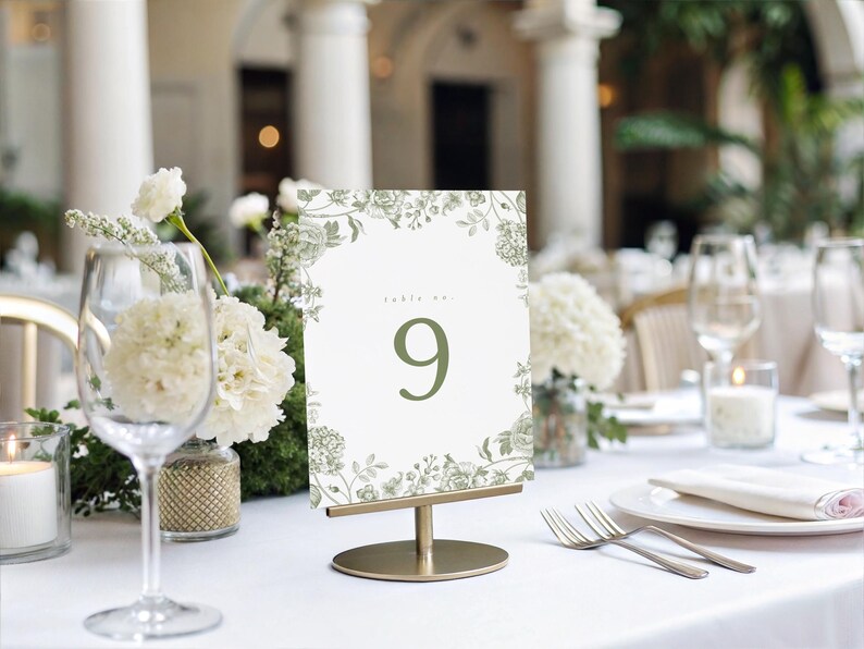 May include: A table setting with a floral table number. The table number is white with a green floral border and the number 9. The table is set with white linens, glassware, and silverware. White flowers and candles are also present.