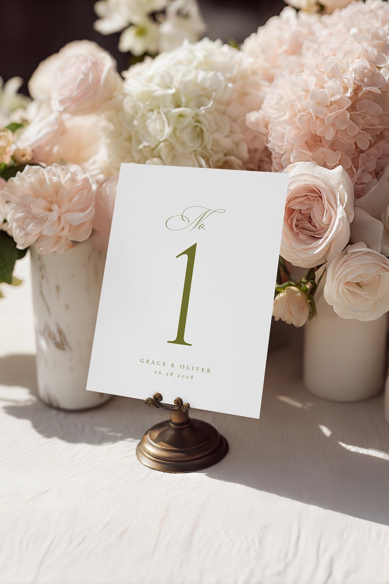 May include: A white table number card with the number "1" in olive green, displayed in a bronze holder. The card also includes the names "Grace & Oliver" and the date "06.28.2028". The card is set against a backdrop of soft pink and white flowers.