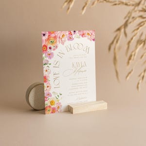May include: A wedding invitation with floral accents and the text "Love is in Bloom." The invitation features a watercolor design with pink, yellow, and purple flowers. It is displayed on a wooden stand.