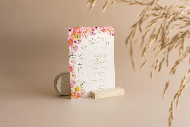 May include: A wedding invitation with floral accents and the text "Love is in Bloom." The invitation features a watercolor design with pink, yellow, and purple flowers. It is displayed on a wooden stand.