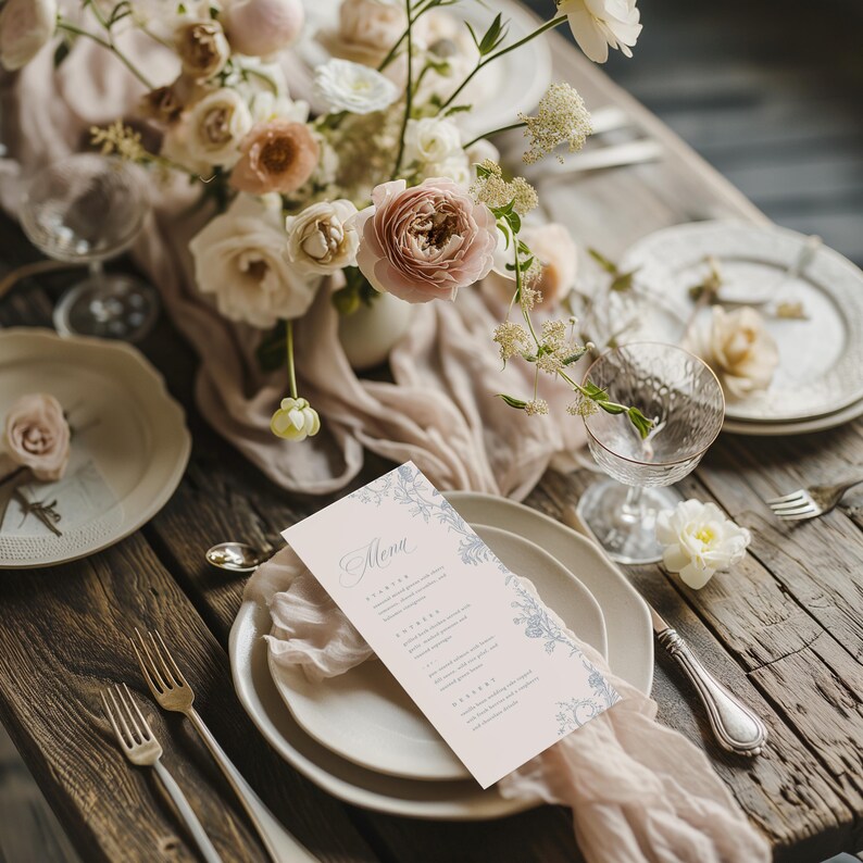May include: A rectangular menu card with a pale pink background and blue floral border accents, featuring the word Menu in script font. The card is placed on a white ceramic plate set on a rustic wooden table with a light pink fabric runner.