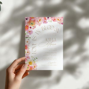 May include: A wedding invitation featuring the words "Love is in Bloom" and the names Kayla and Harrison. The invitation is adorned with pink and white flowers and gold lettering. The invitation is held by a hand.