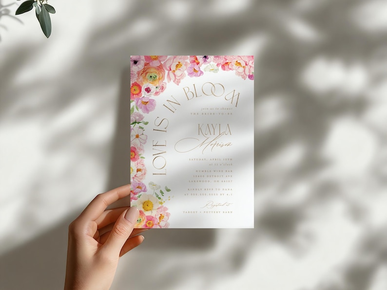 May include: A wedding invitation featuring the words "Love is in Bloom" and the names Kayla and Harrison. The invitation is adorned with pink and white flowers and gold lettering. The invitation is held by a hand.