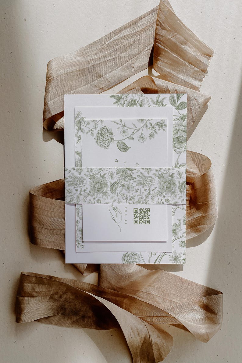May include: A white stationery set featuring green botanical illustrations of flowers and leaves. The set includes a belly band with a floral pattern and a small card with a green QR code and cursive text, all layered on a tan silk ribbon.
