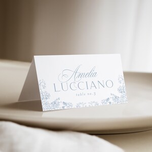 May include: A white folded place card featuring light blue floral illustrations along the bottom edge. The text reads Amelia LUCCIANO and table no. 3 in a combination of elegant script and clean sans-serif typography centered on the front panel.