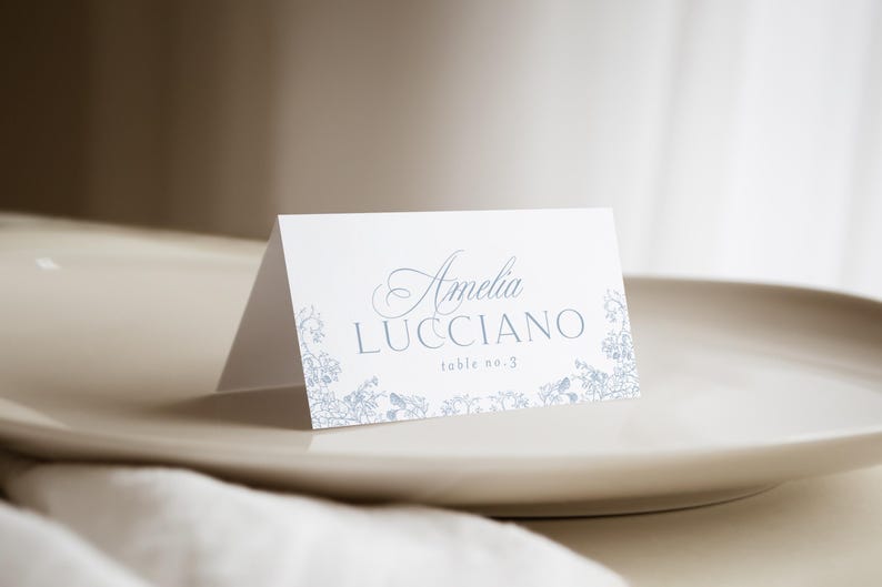 May include: A white folded place card featuring light blue floral illustrations along the bottom edge. The text reads Amelia LUCCIANO and table no. 3 in a combination of elegant script and clean sans-serif typography centered on the front panel.