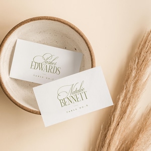 Pode incluir: Dois cart&otilde;es de mesa brancos com letras verdes elegantes, um com a inscri&ccedil;&atilde;o "Parker Edwards" e o outro com "Natalie Bennett". Eles est&atilde;o em uma tigela de cer&acirc;mica salpicada. A grama pampas seca adiciona um toque natural.