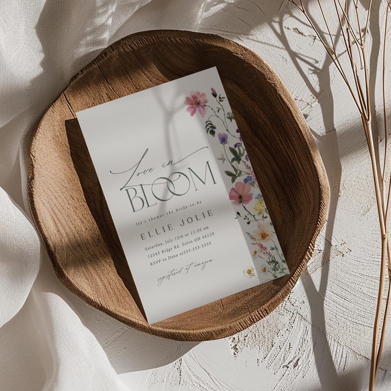 May include: A white invitation card with the text "Love in Bloom" and floral accents. The card includes details for a bridal shower, with the bride's name, date, time, and location. The card is placed in a wooden bowl.